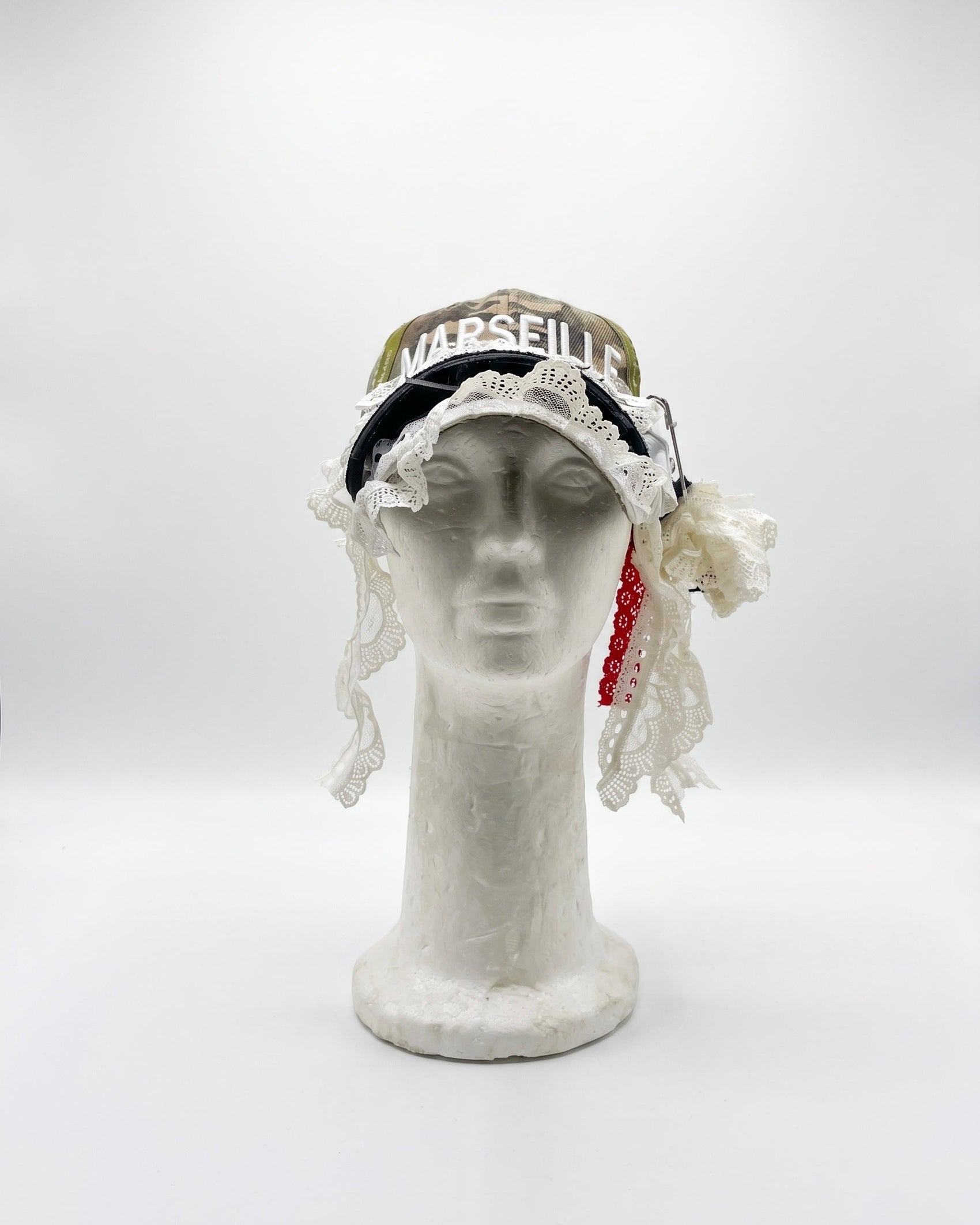 A vintage-style-hat displayed on a white mannequin head against a light gray studio background. The hat features a black band with white lace trim, a gold-toned top section with the word 'Marseille' in white, and a red beaded hanging accent.