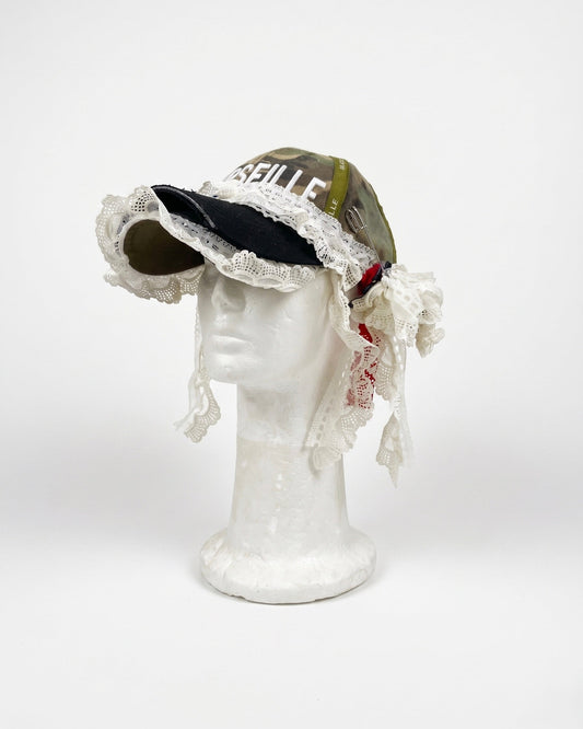 A vintage-style baseball cap displayed on a white mannequin head against a light gray background. The cap features olive green camouflage fabric, a black brim, and prominent white lace trim with dangling ties.